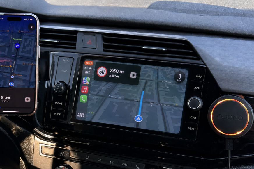 Smartphone zeigt OOONO App mit Navigation und Blitzerwarnung per CarPlay auf Fahrzeugdisplay und Ooono 2 hängt daneben an der Armatur